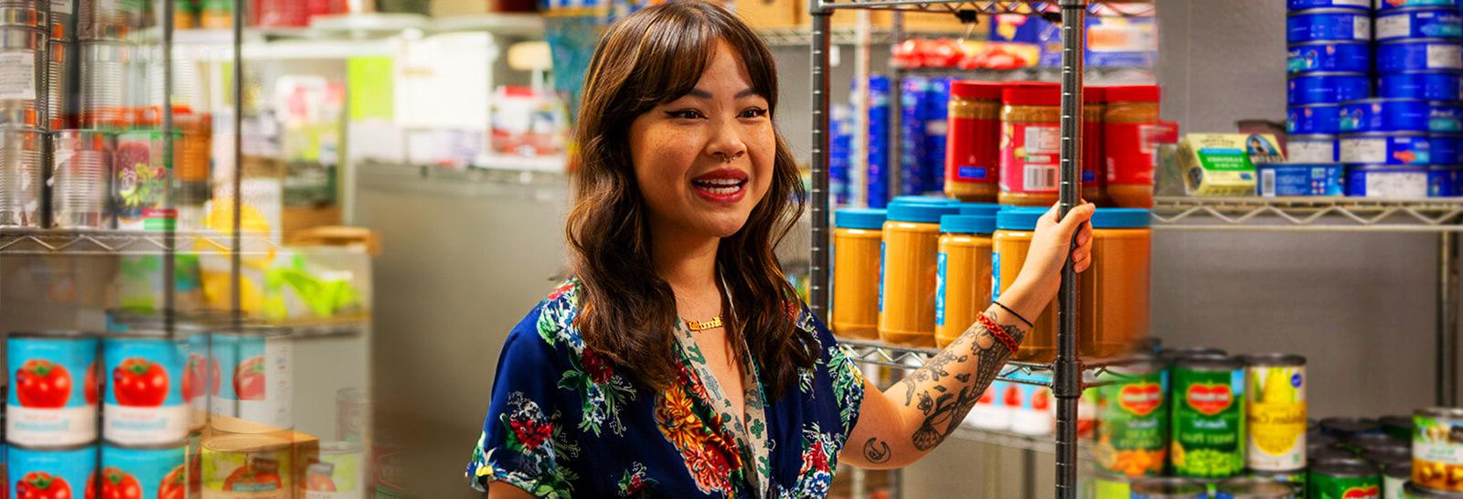 苏珊何 stands next to a shelf of grocery items at the ARC food pantry. 